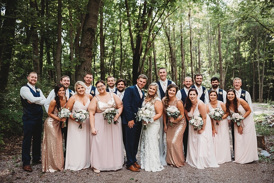 Wedding party with blush dresses and sparkley dresses and navy suits outdoors with a wooded background